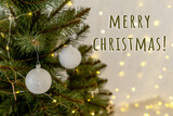 Fototapeta Do pokoju - Part of Christmas tree with white balls, Christmas lights garlands on white brick wall on background. Merry christmas lettering.