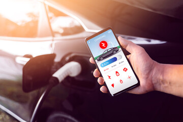 Using mobile smartphone device refueling electric vehicle EV car mock UI application display screen, hand holding cellphone at electric car charging station connected power cable alternative energy