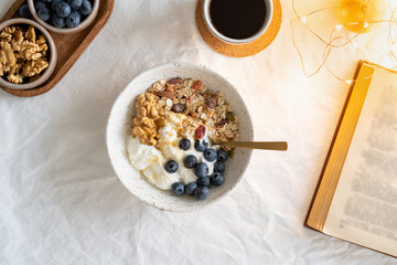 Top view book and Christmas healthy lifestyle breakfast with granola muesli and yogurt in bowl on white table background, cereal grain food with nuts seed. Organic morning diet meal with oat 