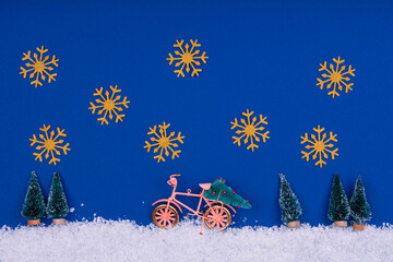 Wall Mural - Funny cozy scene with Christmas toys on classic blue. Christmas tree is moved home by bicycle on the snow road with big golden snowflakes. Horizontal