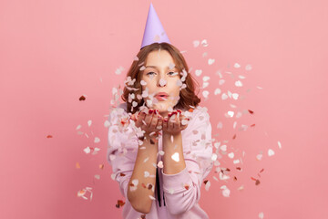 Poster - Portrait of carefree cheerful teenage girl in hoodie and party cone blowing heart shaped confetti, enjoying birthday or valentines day, festive mood. Indoor studio shot, isolated on pink background