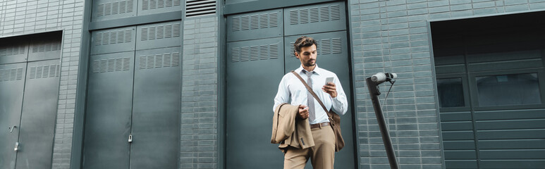 Wall Mural - businessman in suit holding smartphone while standing near e-scooter and building, banner