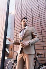 Wall Mural - low angle view of bearded businessman holding smartphone and paper cup near bicycle while standing near building