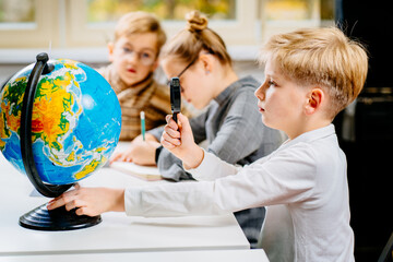 Canvas Print - Smiling boy sits at a table in the classroom and looks at the camera. Portrait of a young little schoolboy studying with classmates in the background.