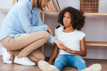 Wall Mural - Psychologist holding hands with upset african american girl with autism sitting on floor near shelves