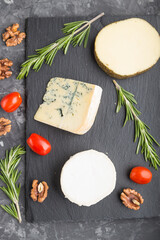 Goat cheese and various types of cheese with rosemary and tomatoes on black slate board on a black concrete background. Top view, close up.