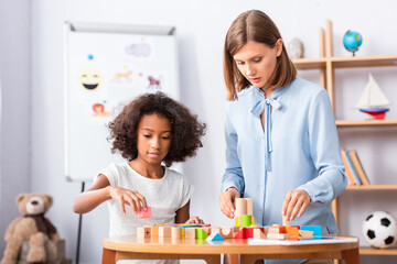 Wall Mural - Psychologist talking to african american girl playing with colorful wooden blocks on coffee table on blurred background