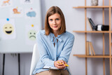 Fototapeta  - Positive female psychologist looking at camera while sitting on chair with blurred office on background
