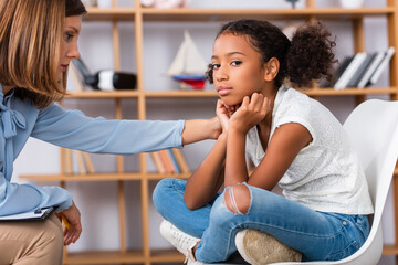 Wall Mural - Psychologist touching shoulder of upset african american girl looking at camera during consultation on blurred background