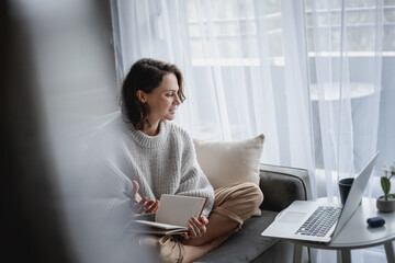 Wall Mural - Beautiful young girl student studying at home online while sitting in a warm sweater on the couch in front of a laptop making notes in a notebook