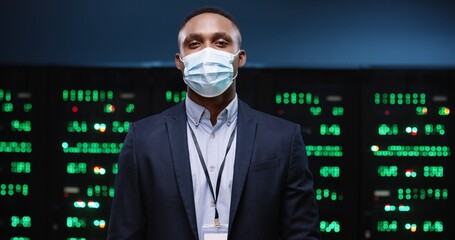 Close up of African American male IT administrator wearing medical mask standing in modern data center with many processors. Portrait of handsome young man security engineer in suit. Business concept