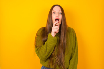 Wall Mural - Nervous puzzled Young beautiful Caucasian woman wearing green sweater against yellow wall opens mouth from surprise, reacts on sudden news.