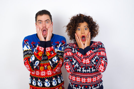 Scared terrified Young couple wearing Christmas sweater standing against white wall shocked with prices at shop, People and human emotions concept