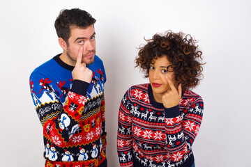 Wall Mural - Young couple wearing Christmas sweater standing against white wall Pointing to the eye watching you gesture, suspicious expression.