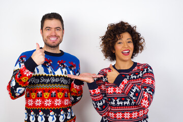 Wall Mural - Young couple wearing Christmas sweater standing against white wall Showing palm hand and doing ok gesture with thumbs up, smiling happy and cheerful.