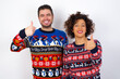 © Roquillo - Young couple wearing Christmas sweater standing against white wall doing happy thumbs up gesture with hand. Approving expression looking at the camera showing success.