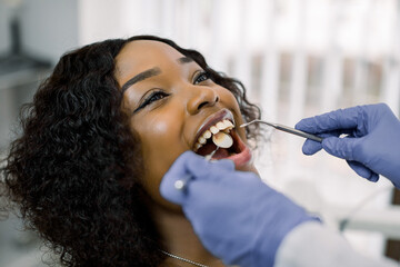 Wall Mural - Close-up portrait of pretty young african woman patient having dental examination and treatment in dentist office, looking at her doctor with smile. Hands of dentist in gloves holding dental tools