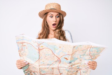 Young caucasian woman with blond hair wearing summer hat holding map in shock face, looking skeptical and sarcastic, surprised with open mouth