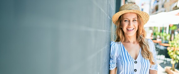 Poster - Young beautiful blonde woman on vacation wearing summer hat smiling happy. Leaning on the wall with smile on face at street of city.