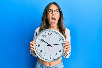 Wall Mural - Young brunette woman holding big clock angry and mad screaming frustrated and furious, shouting with anger looking up.