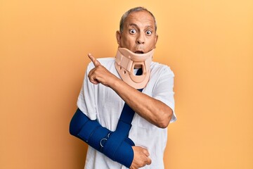 Canvas Print - Handsome mature senior man wearing cervical collar and arm on sling surprised pointing with finger to the side, open mouth amazed expression.