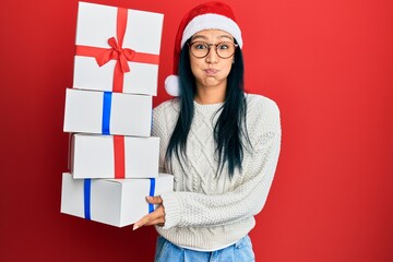 Wall Mural - Beautiful hispanic woman wearing christmas hat and holding gifts puffing cheeks with funny face. mouth inflated with air, catching air.