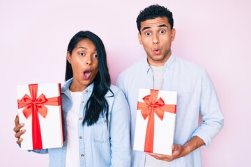 Canvas Print - Beautiful latin young couple holding gift for anniversary celebration scared and amazed with open mouth for surprise, disbelief face