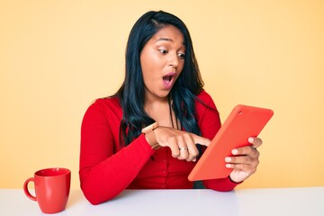 Canvas Print - Beautiful latin young woman with long hair using touchpad sitting on the table scared and amazed with open mouth for surprise, disbelief face