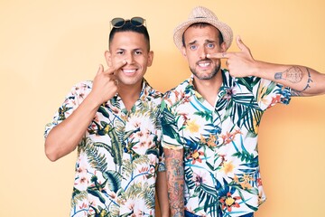 Young gay couple of two men wearing summer hat and hawaiian shirt pointing with hand finger to face and nose, smiling cheerful. beauty concept