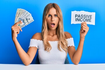 Poster - Beautiful blonde young woman holding dollars and passive income text in shock face, looking skeptical and sarcastic, surprised with open mouth