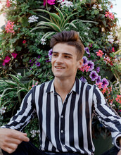 Young Man On A Background Of Flowers. Caucasian Man. Cute Guy Sitting Near Flowers And Plants. A Man Dressed In A Black White Striped Shirt.