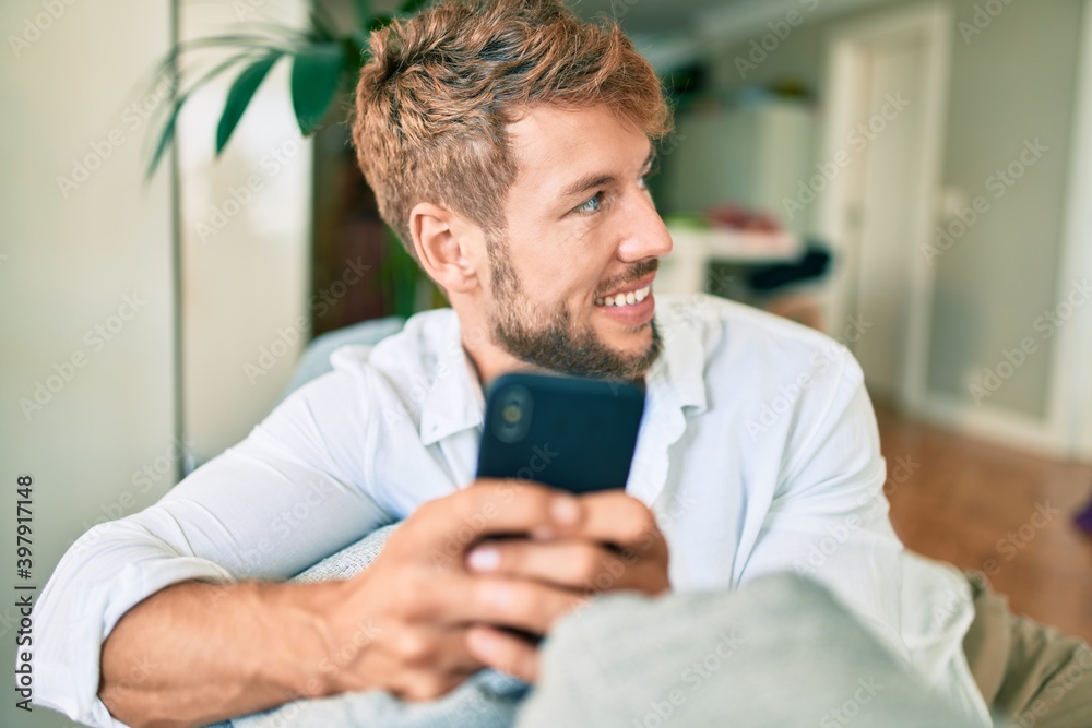 Handsome caucasian man smiling happy sitting on the sofa at home using smartphone - obrazy, fototapety, plakaty 