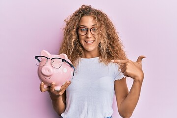 Poster - Beautiful caucasian teenager girl holding piggy bank with glasses pointing finger to one self smiling happy and proud