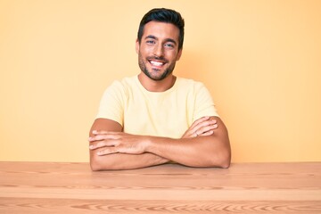 Young hispanic man wearing casual clothes sitting on the table happy face smiling with crossed arms looking at the camera. positive person.