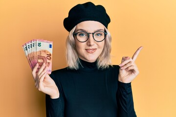 Young blonde girl holding euro banknotes smiling happy pointing with hand and finger to the side