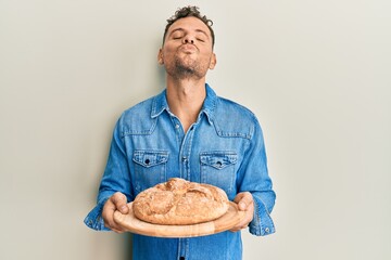 Poster - Young hispanic man holding bread looking at the camera blowing a kiss being lovely and sexy. love expression.