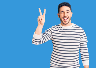 Young hispanic man wearing casual clothes smiling with happy face winking at the camera doing victory sign with fingers. number two.