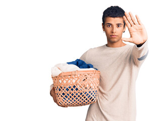 Poster - Young african amercian man holding laundry basket with open hand doing stop sign with serious and confident expression, defense gesture