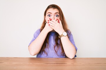 Wall Mural - Young redhead woman wearing casual clothes sitting on the table shocked covering mouth with hands for mistake. secret concept.