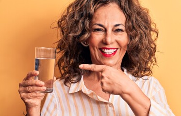 Middle age brunette woman drinking glass of water standing over isolated yellow background smiling happy pointing with hand and finger