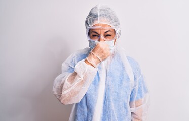 Wall Mural - Middle age nurse woman wearing protection coronavirus equipment over white background smelling something stinky and disgusting, intolerable smell, holding breath with fingers on nose. Bad smell