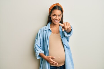 Sticker - Beautiful hispanic woman expecting a baby, touching pregnant belly pointing to you and the camera with fingers, smiling positive and cheerful