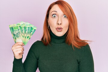 Poster - Beautiful redhead woman holding south african 10 rand banknotes scared and amazed with open mouth for surprise, disbelief face