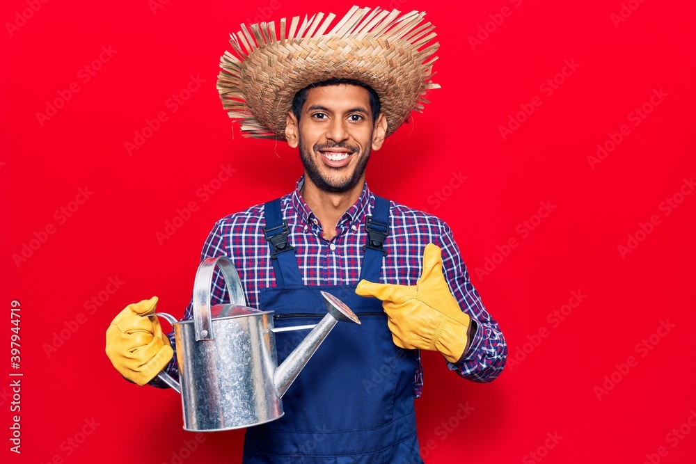 Young latin man wearing farmer hat and gloves holding watering can smiling happy pointing with hand and finger