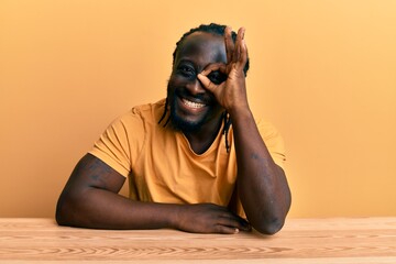 Sticker - Handsome young black man wearing casual clothes sitting on the table doing ok gesture with hand smiling, eye looking through fingers with happy face.