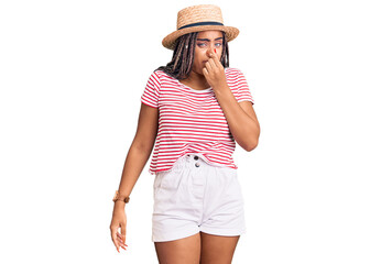 Sticker - Young african american woman with braids wearing summer hat smelling something stinky and disgusting, intolerable smell, holding breath with fingers on nose. bad smell