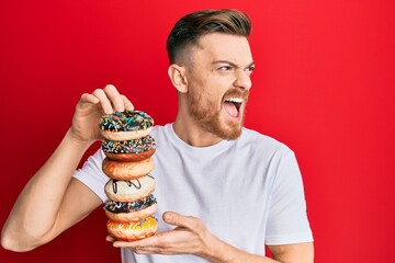 Wall Mural - Young redhead man holding pile of tasty colorful doughnuts angry and mad screaming frustrated and furious, shouting with anger. rage and aggressive concept.