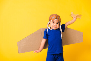 Wall Mural - Happy Asian handsome funny child or kid little boy smile wear pilot hat play and goggles with toy cardboard airplane wings fly hold plane toy, studio shot isolated yellow background, Startup freedom