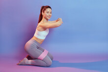 Photo Of Joyful Active Sportive Girl Doing Sport Exercise, Sitting On Knees With Pink Expander, Looking Smiling At Camera, Posing Isolated Over Blue Background.
