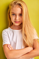 Wall Mural - nice caucasian little girl with long blond hair isolated on green background, adorable girl looks at camera holding arms crossed